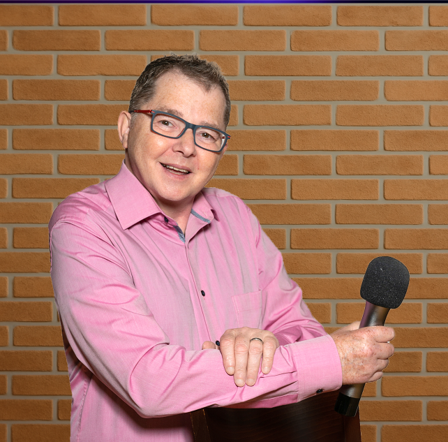 Stefan Fess in rosa Hemd mit Mikrofon, lächelnd vor einer Ziegelwand - Hochzeits-DJ im Saarland